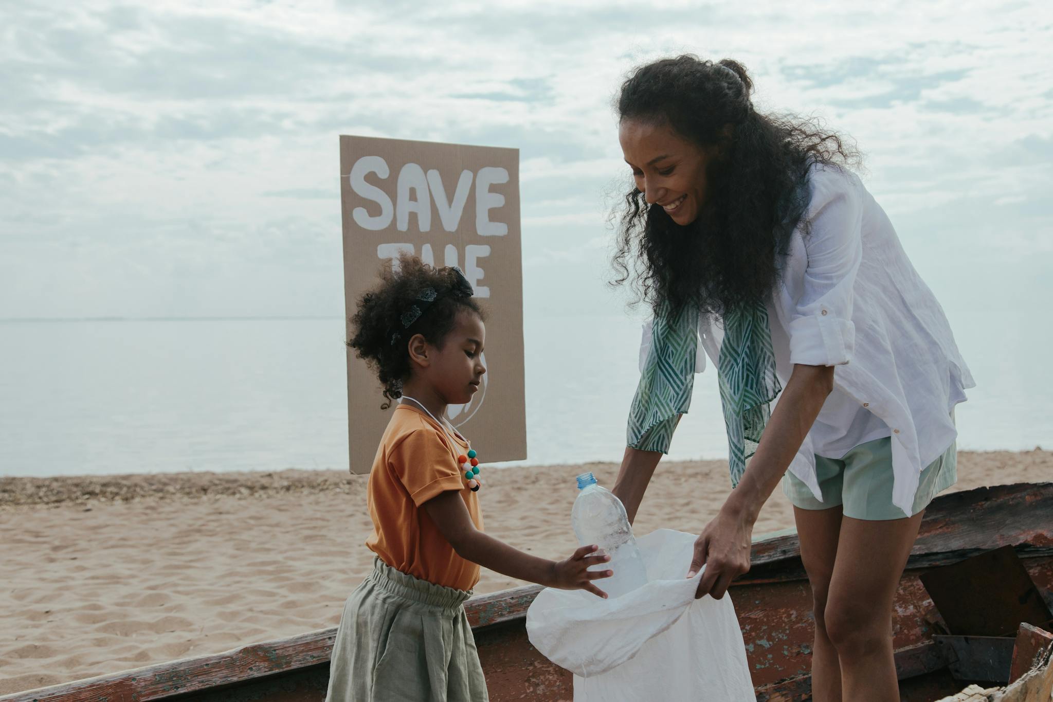 Mother and daughter cleaning beach, promoting environment protection with a 'Save the Ocean' sign.